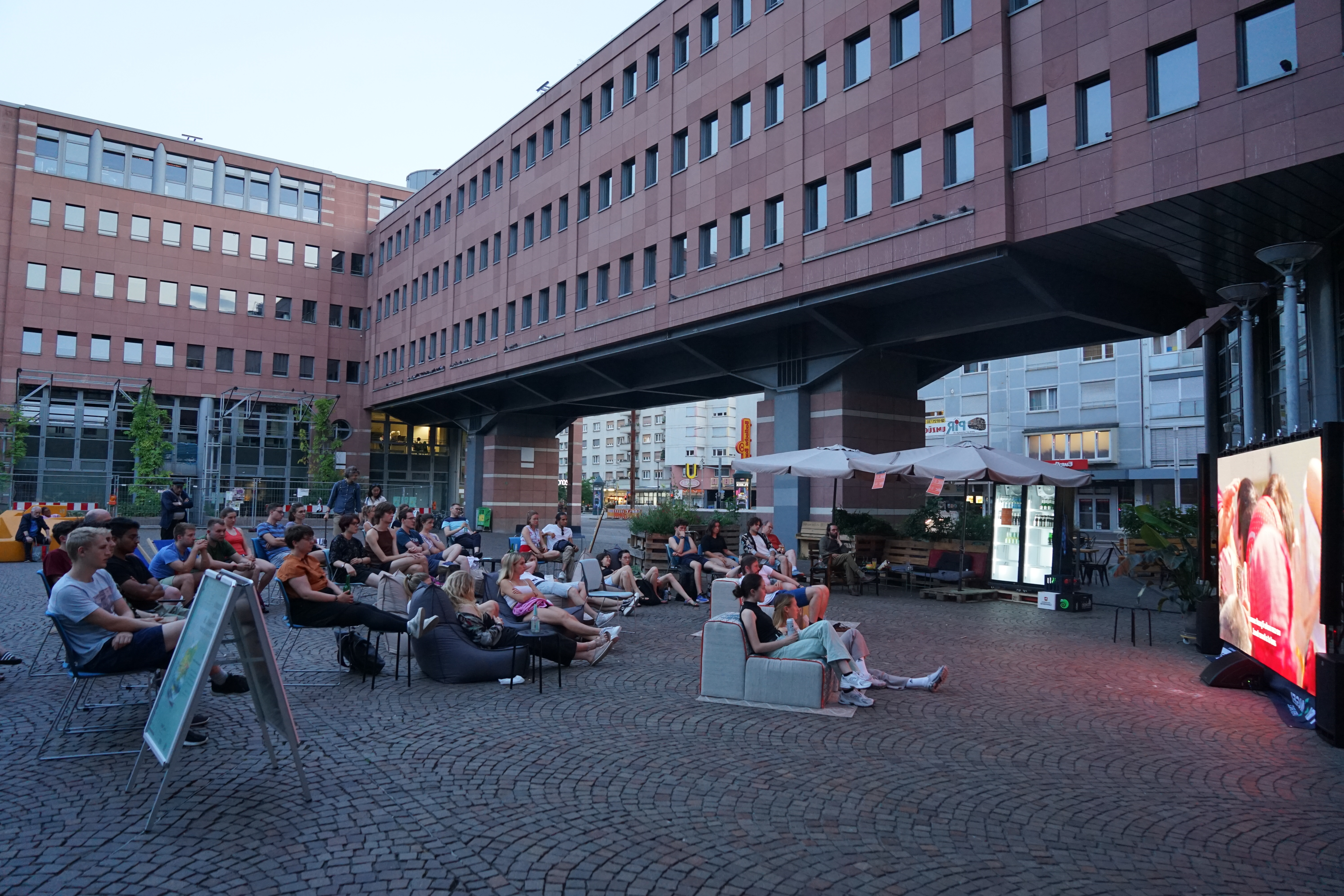 Open-Air-Kino, eine Gruppe von Menschen schaut auf eine große Leinwand auf der rechten Bildseite